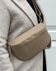 woman wearing a large light taupe crossbody/ waist bag  with and adjustable strap and gold hardware. 